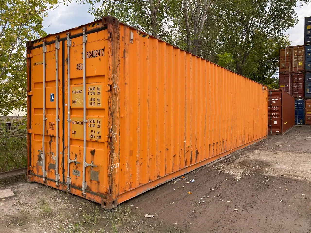 Used orange shipping container with dents, rust, and graffiti at a depot