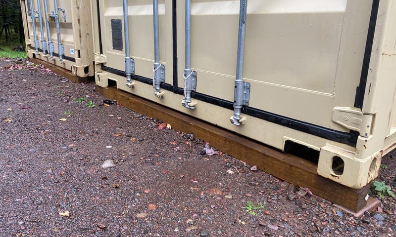 Two shipping containers sitting on wooden beams over gravel, with the door frames supported off the ground.